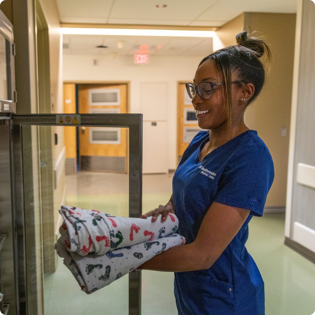 A smiling nurse in blue scrubs is taking a stack of folded blankets out of a warming cabinet in a hallway