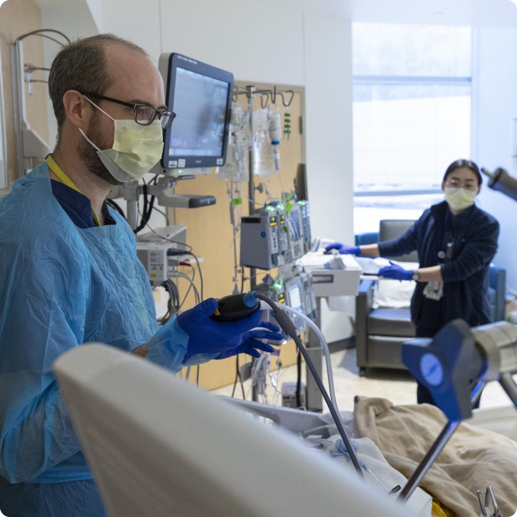 A male nurse in a mask, gown and gloves working with medical equipment in an ICU patient room.