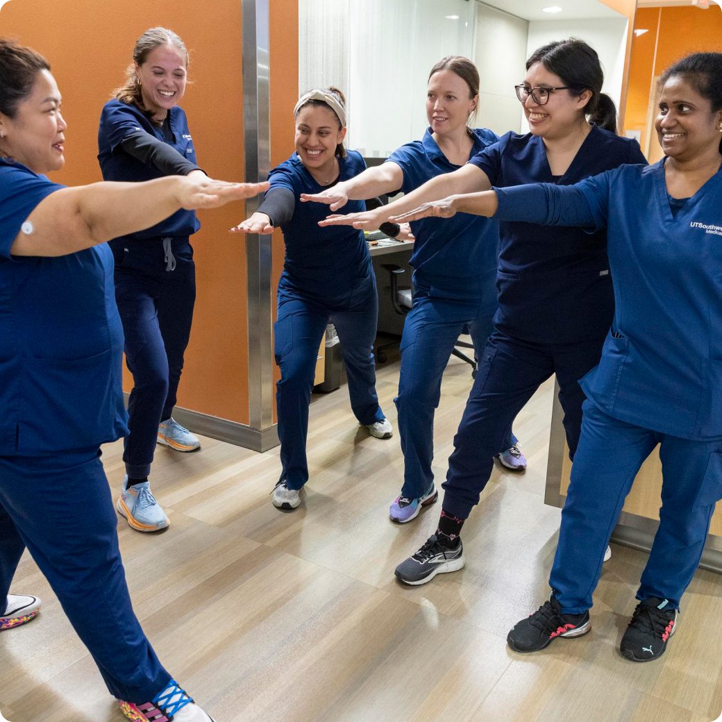 Six female nurses in blue scrubs having a team huddle with their hands in the middle in the hospital hallway.