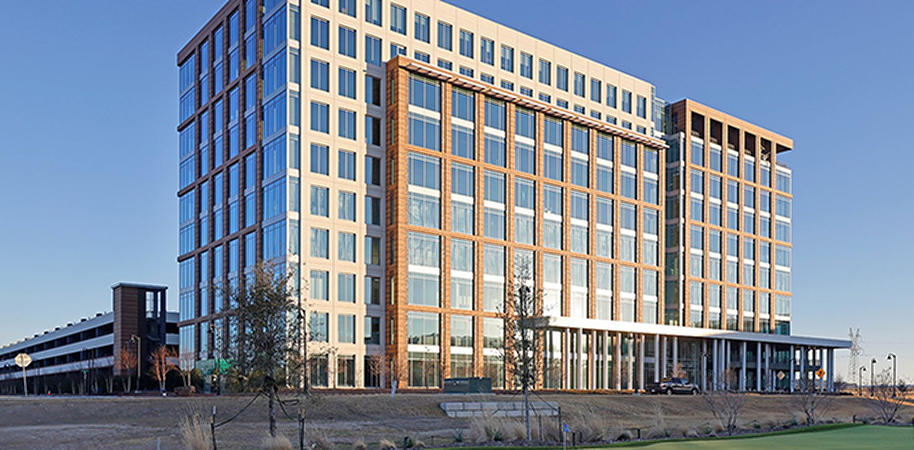 UT Southwestern Coppell, a modern glass office building with brown accents, surrounded by open land, trees, and a parking garage under a clear blue sky.