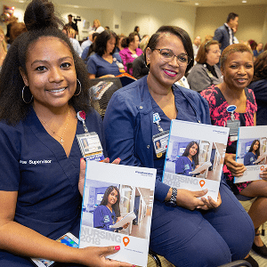 Three nurses sit together at an event, smiling and holding nursing reports, with other attendees seated in the background.